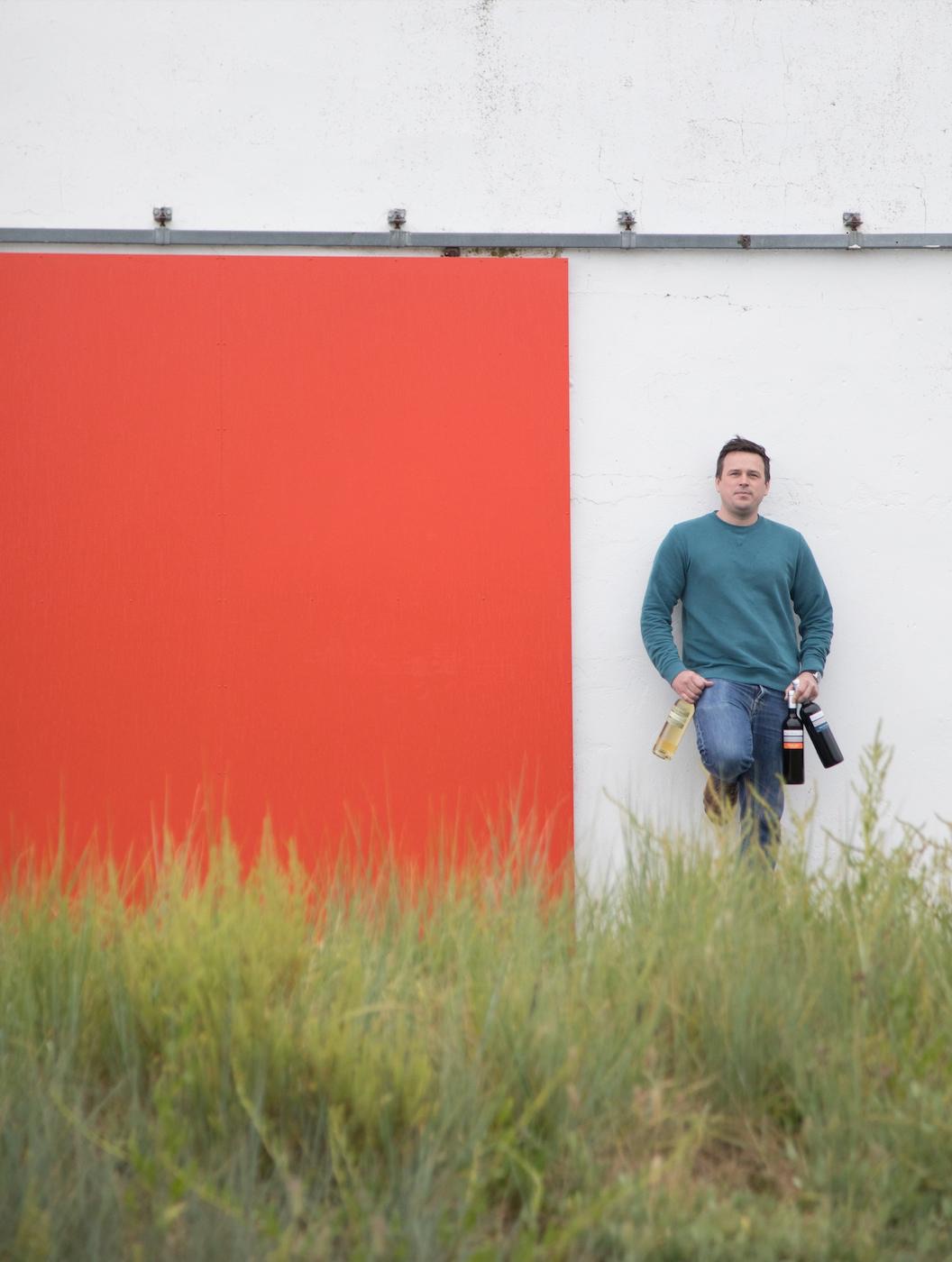 Cyrille Cornerotte, vigneron de Pique Russe, adossé à un mur rouge avec ses bouteilles
