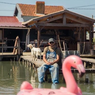 La Cabane Buissonnière — Manuel assis sur un ponton, avec un flamant rose gonflable au premier plan et une cabane ostréicole en arrière-plan
