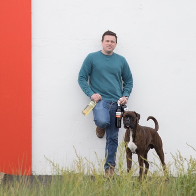 Cyrille Cornerotte, vigneron artisan, adossé à un mur blanc avec ses bouteilles et son chien