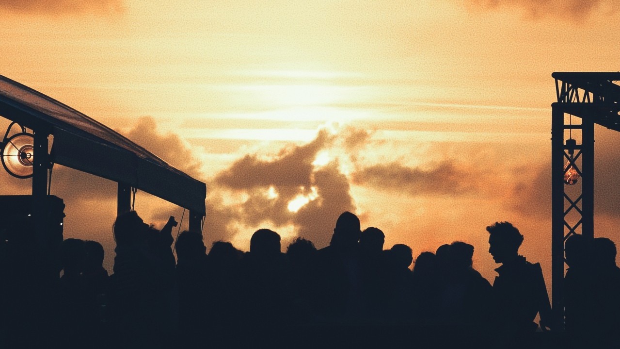 Silhouettes de convives devant un ciel de coucher de soleil orangé, structure de scène et éclairage